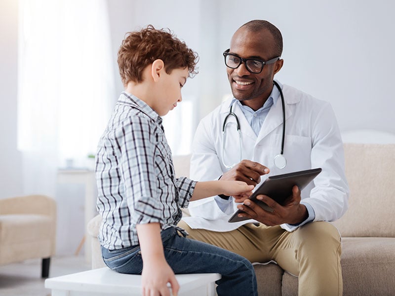 Positive-pleasant-male-doctor-smiling-while-showing-tablet-to-boy-and-sitting
