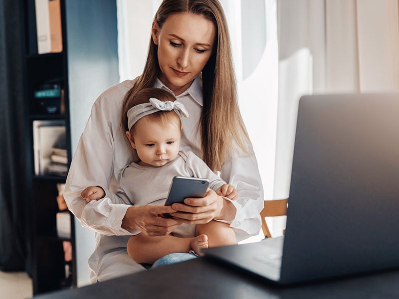 Young-woman-texting-on-smartphone-with-baby-girl-sitting-on-her-knees-and-looking-at-screen
